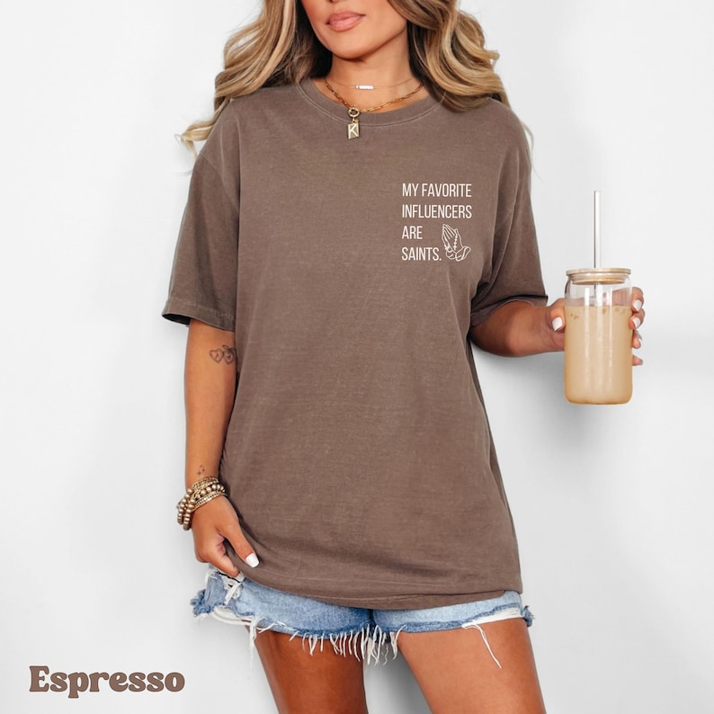 A model wears an espresso color shirt with a pockey-size design over the upper left chest (viewed on the right when looking at the model). It says in all capital in a modern san serif font separated into 4 lines, "My favorite influencers are Saints." There are prayer hands next to it grasping a rosary with a cross hanging on beads.