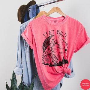 May include: A pink t-shirt with a black wolf graphic and the text "STAY WILD" printed on the front. The wolf is howling at the moon, which is behind it. The wolf is surrounded by flowers and trees.