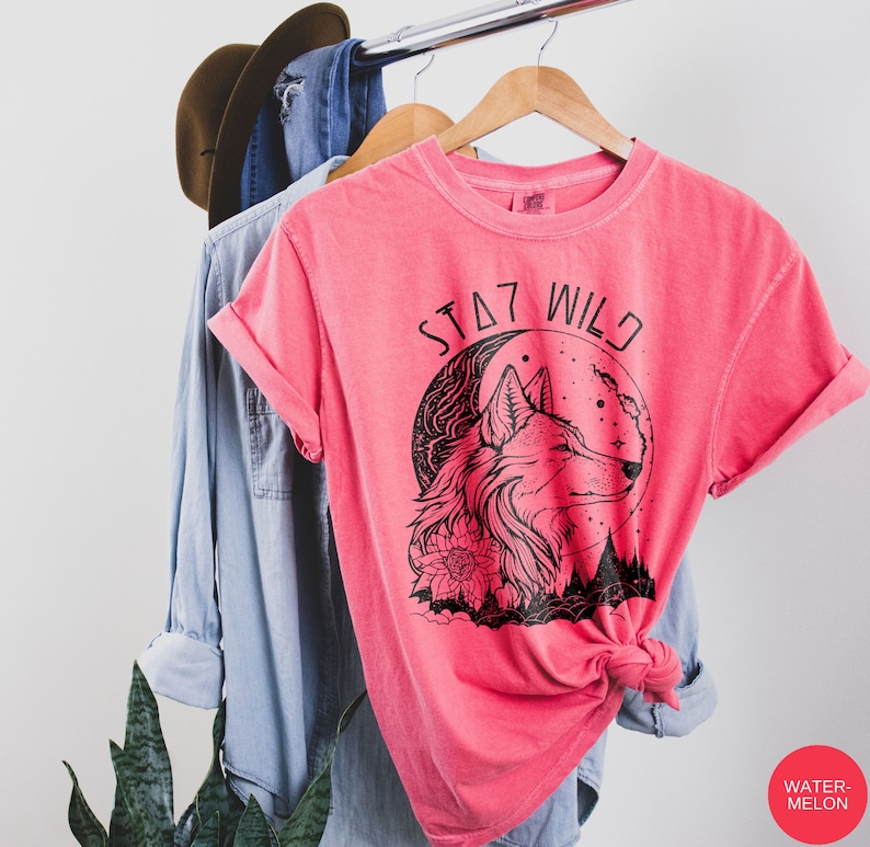 May include: A pink t-shirt with a black wolf graphic and the text "STAY WILD" printed on the front. The wolf is howling at the moon, which is behind it. The wolf is surrounded by flowers and trees.
