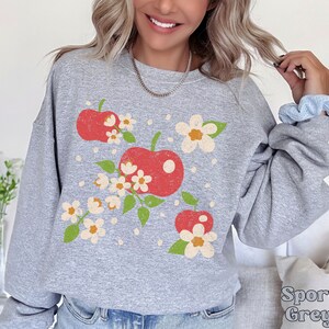 May include: A gray sweatshirt with a vintage-style graphic of red apples and white flowers. The sweatshirt has a crew neck and long sleeves.