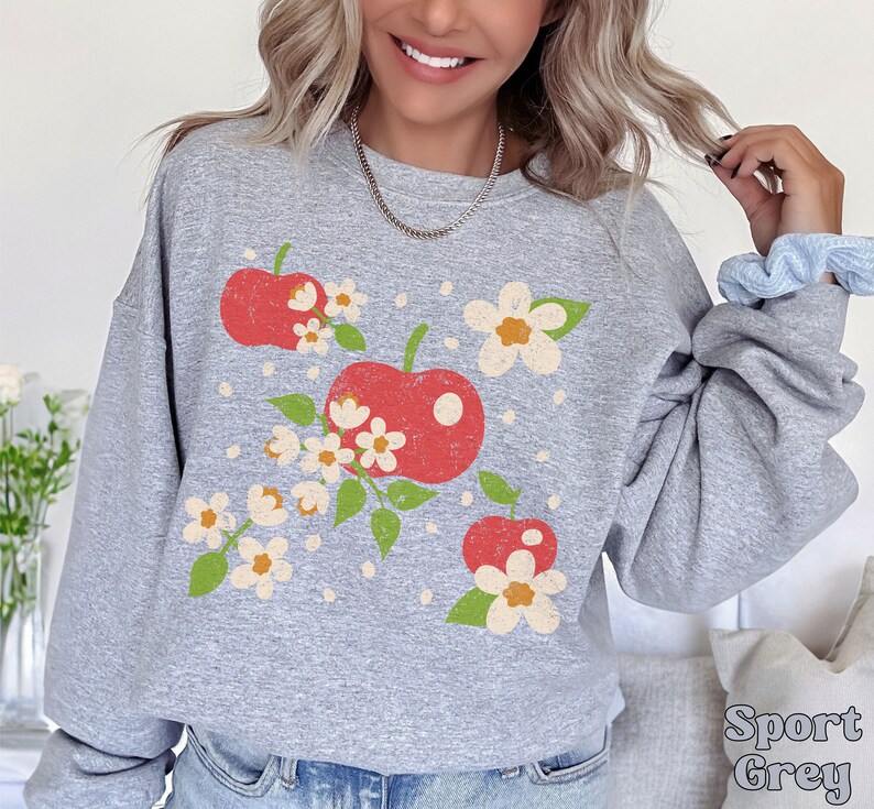 May include: A gray sweatshirt with a vintage-style graphic of red apples and white flowers. The sweatshirt has a crew neck and long sleeves.