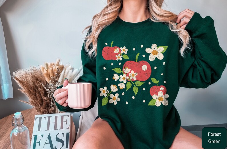 May include: A forest green sweatshirt with a vintage-style graphic print of red apples and white flowers.