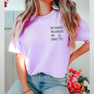 May include: Lilac t-shirt featuring the text "MY FAVORITE INFLUENCERS ARE SAINTS" and a praying hands graphic. The shirt is a solid colour and has short sleeves. The word "Orchid" is printed at the bottom left of the image.