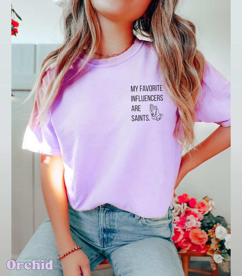 May include: Lilac t-shirt featuring the text "MY FAVORITE INFLUENCERS ARE SAINTS" and a praying hands graphic. The shirt is a solid colour and has short sleeves. The word "Orchid" is printed at the bottom left of the image.