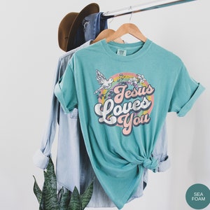 May include: A teal blue t-shirt with a retro-style graphic that says "Jesus Loves You" in a rainbow-colored circle with a white dove and flowers.