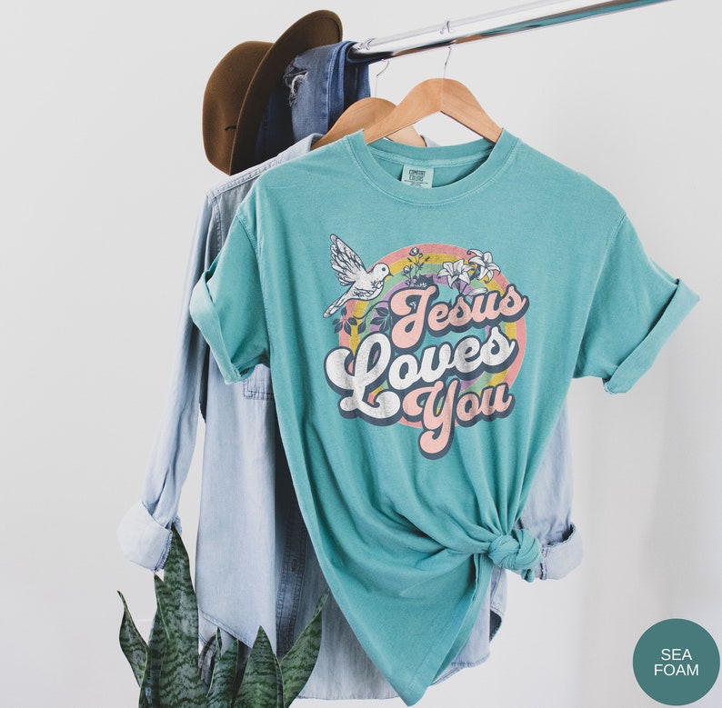 May include: A teal blue t-shirt with a retro-style graphic that says "Jesus Loves You" in a rainbow-colored circle with a white dove and flowers.