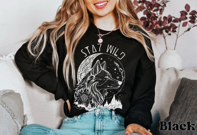 A model wears a black sweatshirt that says Stay Wild in a futuristic arch and a boho graphic in white outline of a profile of Wolf in front of a crescent moon with stars. There are trees in the distance. The wolf has some flowers on their neck.