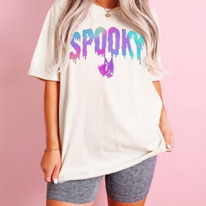 May include: Ivory-colored t-shirt with the word "SPOOKY" in a dripping, multi-colored font. A bat graphic is below the word. The shirt is paired with gray shorts. The word "Ivory" is printed at the bottom of the image.
