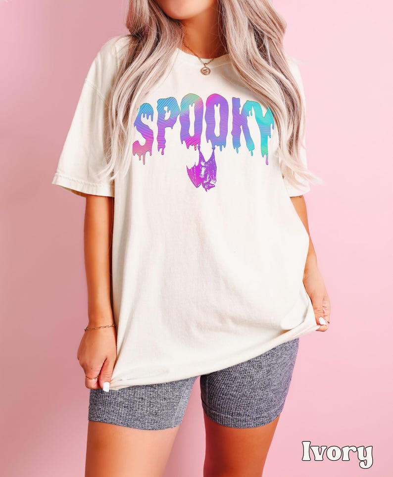 May include: Ivory-colored t-shirt with the word "SPOOKY" in a dripping, multi-colored font. A bat graphic is below the word. The shirt is paired with gray shorts. The word "Ivory" is printed at the bottom of the image.