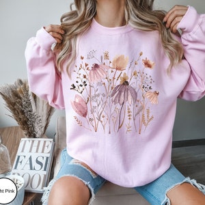 May include: A light pink sweatshirt with a watercolor floral print featuring pink, brown, and white flowers. The sweatshirt has long sleeves and a crew neck.