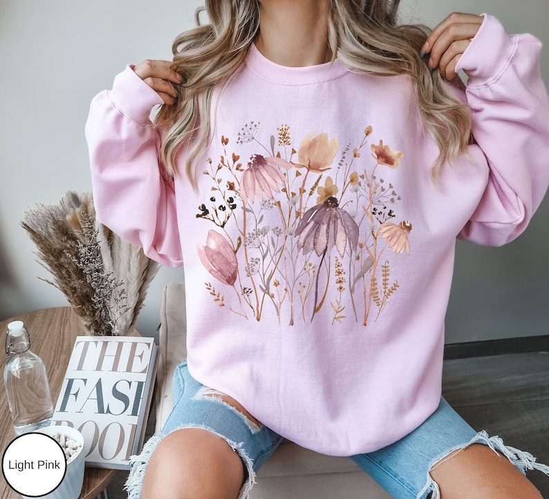 May include: A light pink sweatshirt with a watercolor floral print featuring pink, brown, and white flowers. The sweatshirt has long sleeves and a crew neck.