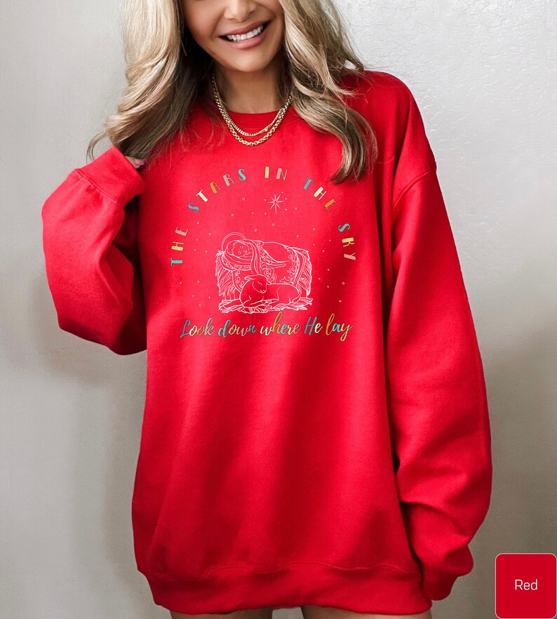 May include: Red sweatshirt with a Christmas-themed design featuring a nativity scene and the text "The stars in the sky look down where He lay."