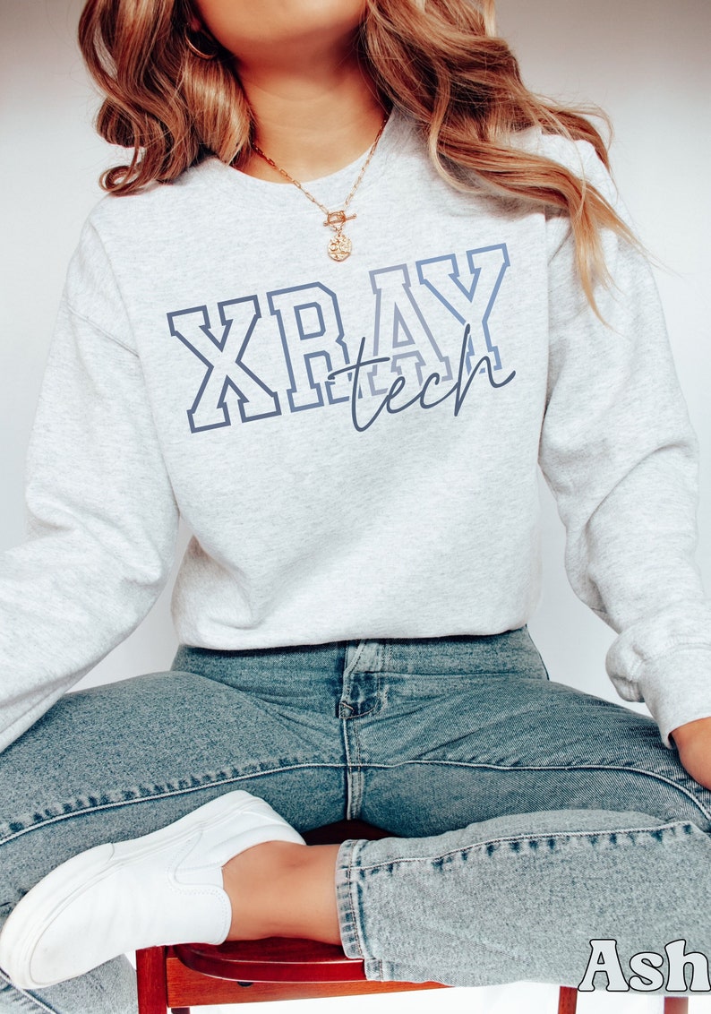 A model wears an ash colored sweatshirt that says Xray tech. Xray is written in a large blocky outline font in different shades of blue from dark to light-gray-blue. Tech is written in smaller lowercase script in the lower right of xray.