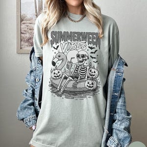 May include: A light green t-shirt with the text "Summerween Vibes" and a graphic of a skeleton relaxing on a flamingo floatie, wearing sunglasses, and surrounded by jack-o'-lanterns and bats.