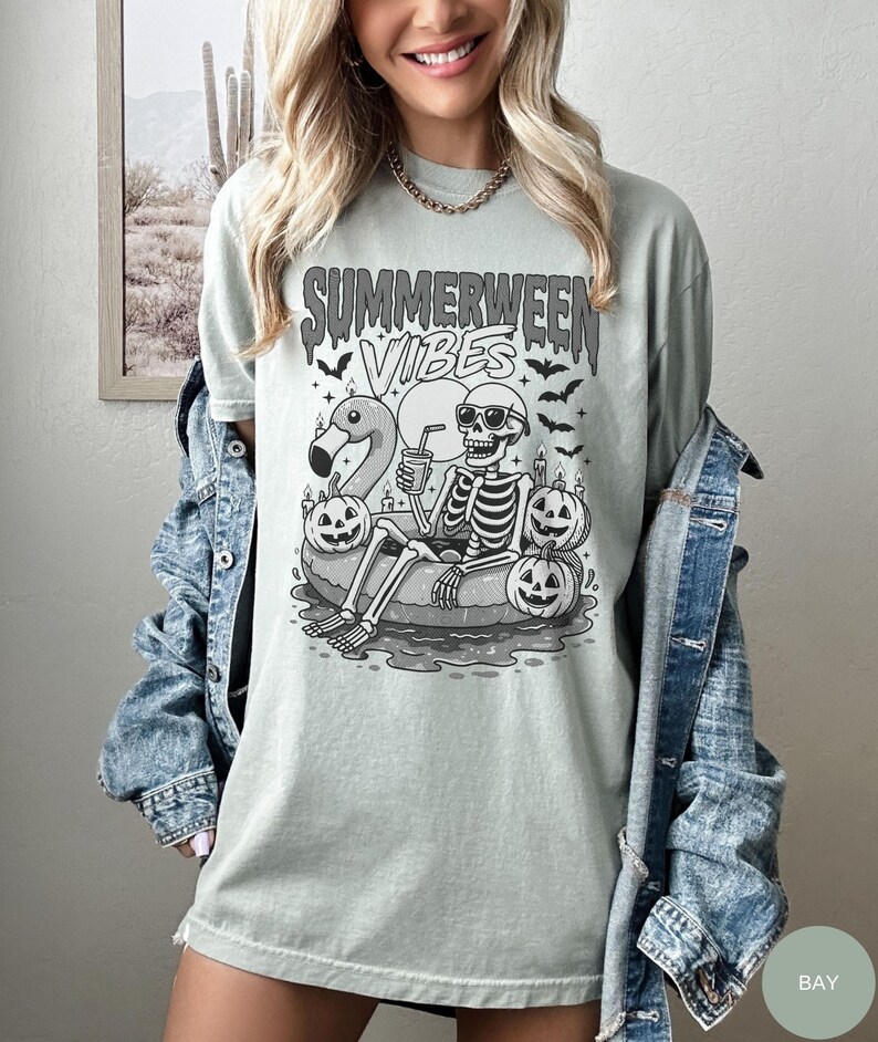 May include: A light green t-shirt with the text "Summerween Vibes" and a graphic of a skeleton relaxing on a flamingo floatie, wearing sunglasses, and surrounded by jack-o'-lanterns and bats.