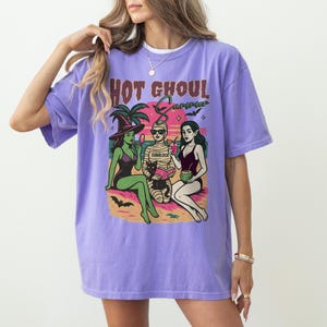 May include: A lilac-coloured t-shirt featuring the text "Hot Ghoul Summer" above a graphic of three cartoon figures in swimwear. The graphic depicts a witch, a mummy, and a vampire on a beach. The t-shirt has a loose fit.