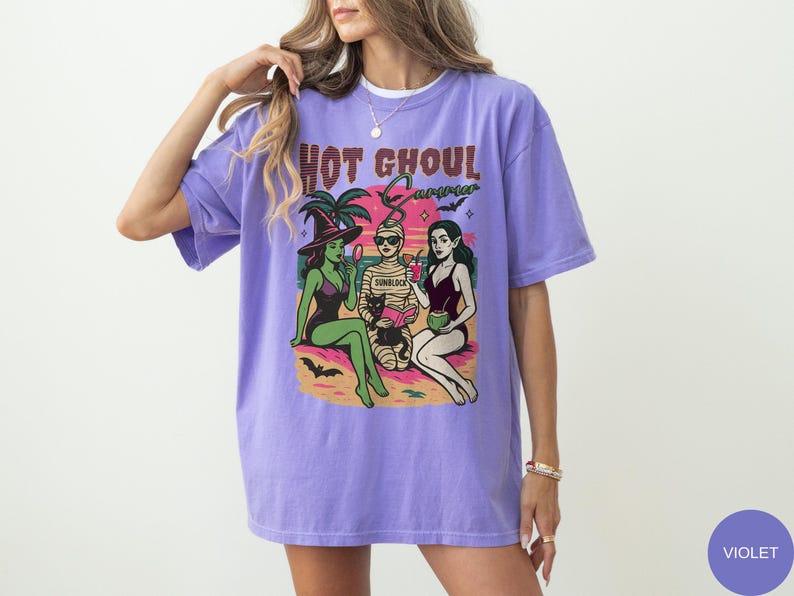 May include: A lilac-coloured t-shirt featuring the text "Hot Ghoul Summer" above a graphic of three cartoon figures in swimwear. The graphic depicts a witch, a mummy, and a vampire on a beach. The t-shirt has a loose fit.
