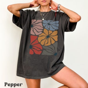 A model wears a pepper shirt with abstract, wavy, boho flowers in colors of muted mauve, light grey-blue, gold, red, and dark taupe all with deep blue pistils. The colorful design is a slightly distressed texture.