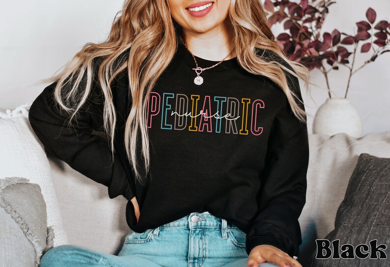 May include: A black sweatshirt with the text "PEDIATRIC nurse" in rainbow colors. The text is outlined in black. The model is wearing blue jeans.