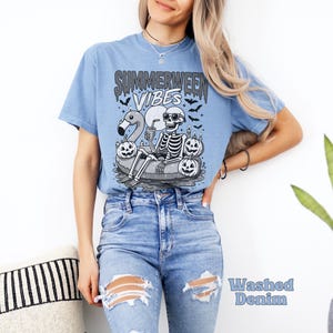 May include: Light blue t-shirt with a graphic of a skeleton in sunglasses, floating on an inner tube with a flamingo, pumpkins, and the text "Summerween Vibes." The shirt is made of a soft material.