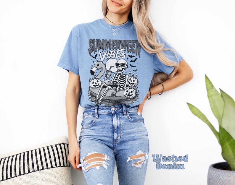 May include: Light blue t-shirt with a graphic of a skeleton in sunglasses, floating on an inner tube with a flamingo, pumpkins, and the text "Summerween Vibes." The shirt is made of a soft material.