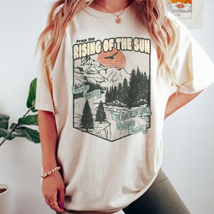 May include: A beige t-shirt featuring a vintage-style graphic of a mountain landscape and a sunset. The graphic includes the text "From the Rising of the Sun" and a bible verse, "The name of the Lord is to be praised, Psalm 113:3".