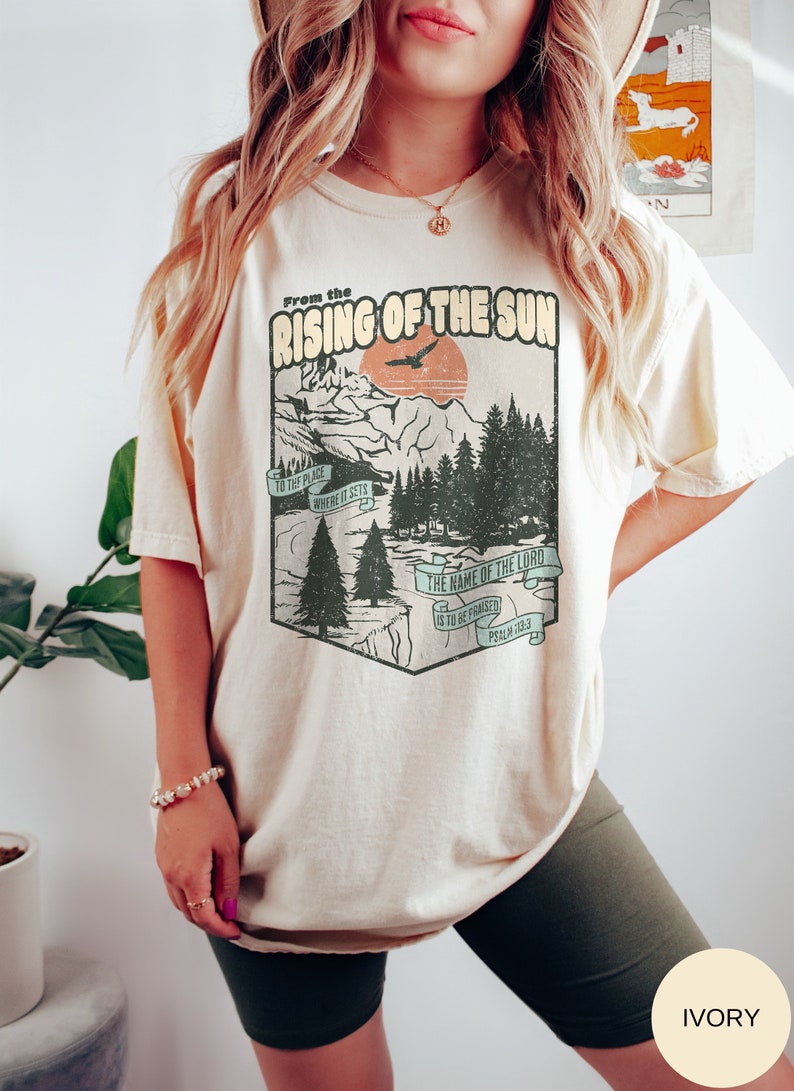 May include: A beige t-shirt featuring a vintage-style graphic of a mountain landscape and a sunset. The graphic includes the text "From the Rising of the Sun" and a bible verse, "The name of the Lord is to be praised, Psalm 113:3".