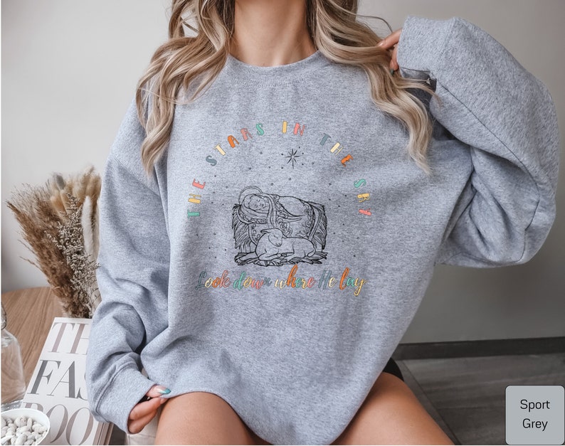 May include: A gray sweatshirt with a colorful graphic design that says "The stars in the sky look down where he lay". The graphic features a star, a manger, and a baby.