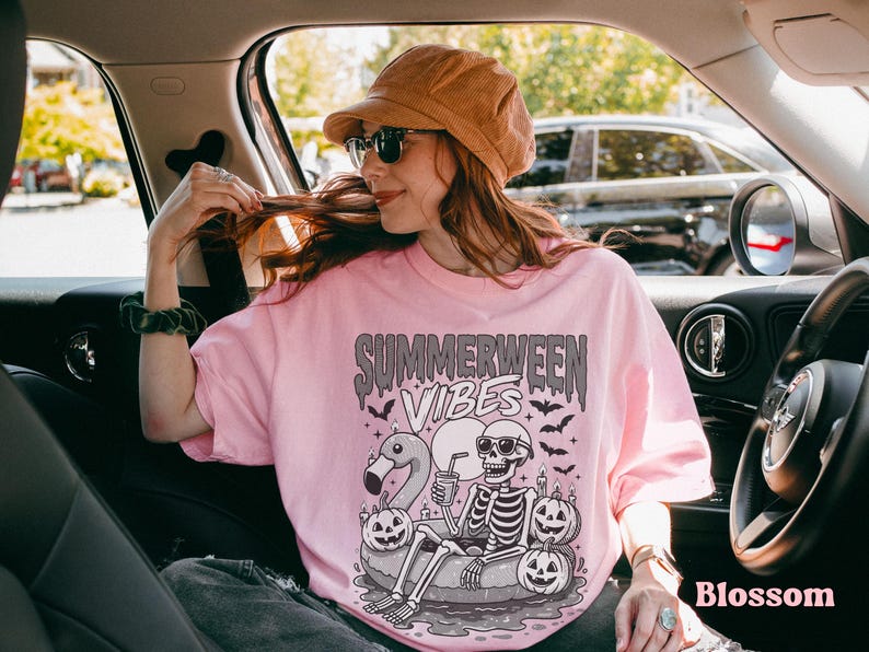 May include: A light pink t-shirt with the text "Summerween Vibes" in a spooky font. The graphic features a skeleton in sunglasses on a float with pumpkins and a flamingo. The model is wearing a corduroy hat and sunglasses.
