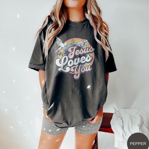 May include: A black t-shirt with a retro-style graphic that says "Jesus Loves You" in a rainbow circle with a white dove and flowers.