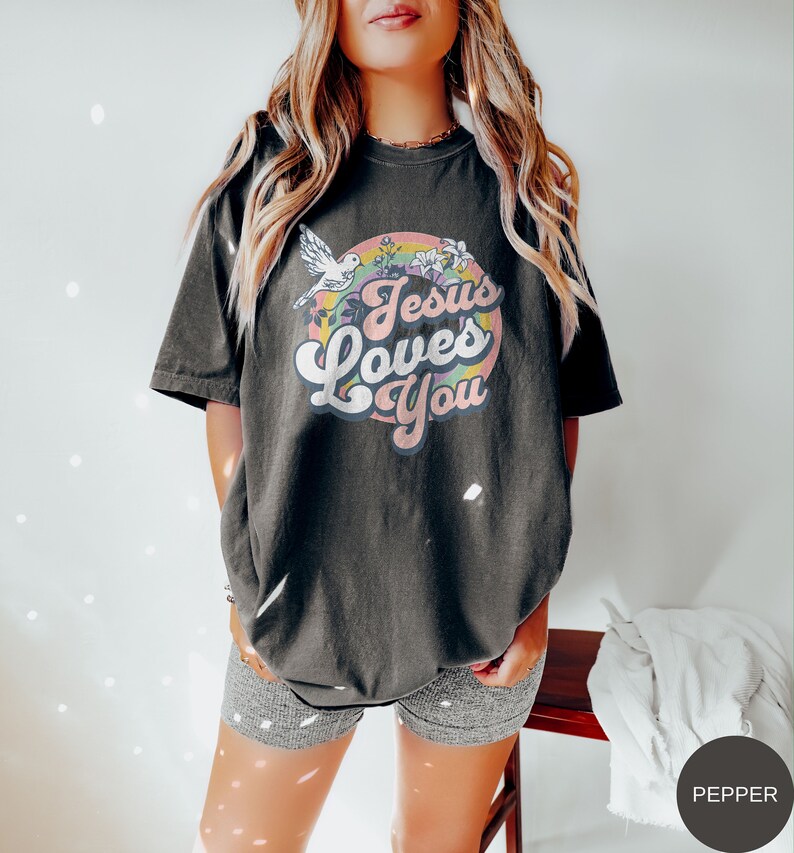 May include: A black t-shirt with a retro-style graphic that says "Jesus Loves You" in a rainbow circle with a white dove and flowers.