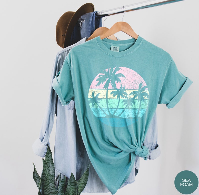 以下が含まれることがあります： パステルカラーの夕焼けとヤシの木のヴィンテージスタイルのグラフィックがプリントされたターコイズブルーのTシャツ。画像の右下隅に「SEA FOAM」の文字が見えます。