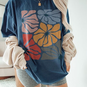 A model wears a midnight shirt with abstract, wavy, boho flowers in colors of muted mauve, light grey-blue, gold, red, and dark taupe all with deep blue pistils. The colorful design is a slightly distressed texture.