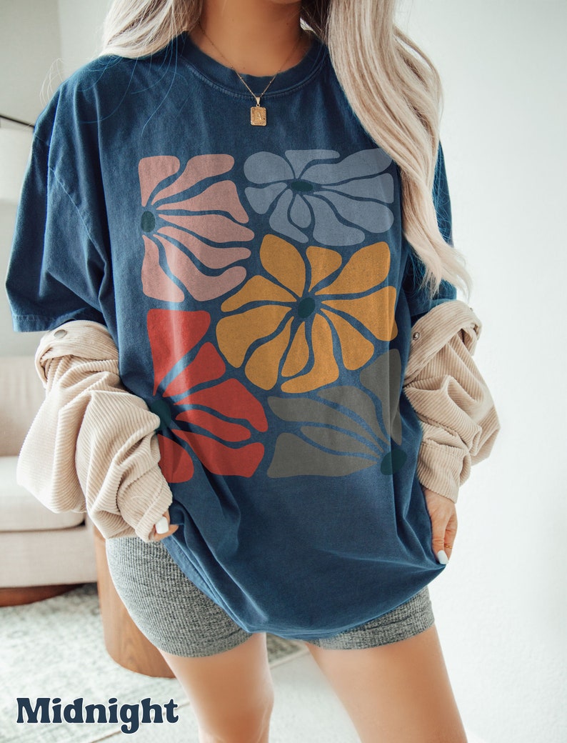 A model wears a midnight shirt with abstract, wavy, boho flowers in colors of muted mauve, light grey-blue, gold, red, and dark taupe all with deep blue pistils. The colorful design is a slightly distressed texture.