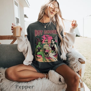 May include: Dark grey t-shirt featuring a graphic with the text "Hot Ghoul Summer" above a beach scene with three cartoon figures. The figures are a witch, a mummy, and a vampire. The witch is wearing a purple swimsuit and a black hat. The mummy is wearing sunglasses.