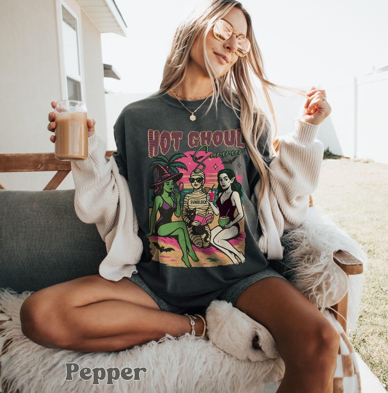 May include: Dark grey t-shirt featuring a graphic with the text "Hot Ghoul Summer" above a beach scene with three cartoon figures. The figures are a witch, a mummy, and a vampire. The witch is wearing a purple swimsuit and a black hat. The mummy is wearing sunglasses.