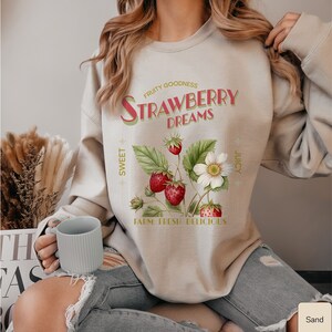 May include: A beige sweatshirt with a vintage-style graphic of strawberries and the text "FRUITY GOODNESS STRAWBERRY DREAMS SWEET JUICY FARM FRESH DELICIOUS".