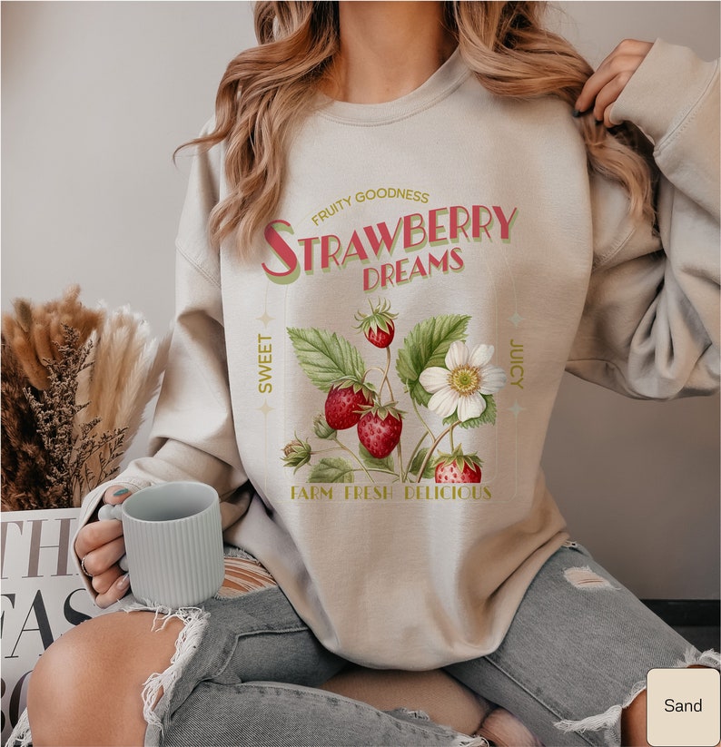 May include: A beige sweatshirt with a vintage-style graphic of strawberries and the text "FRUITY GOODNESS STRAWBERRY DREAMS SWEET JUICY FARM FRESH DELICIOUS".