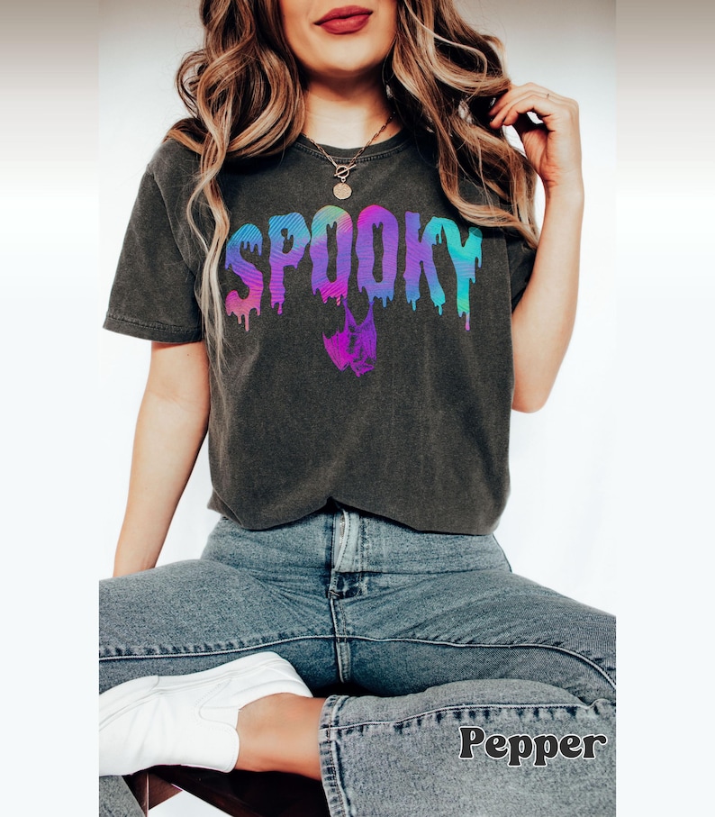 A model wears a pepper color shirt that says Spooky in an arched drippy font with a hanging bat hanging below the 2 Os. The colors are a pastel-like rainbow of iridescent colors gradient from one corner to the other. It has a retro, preppy, holographic vibe.