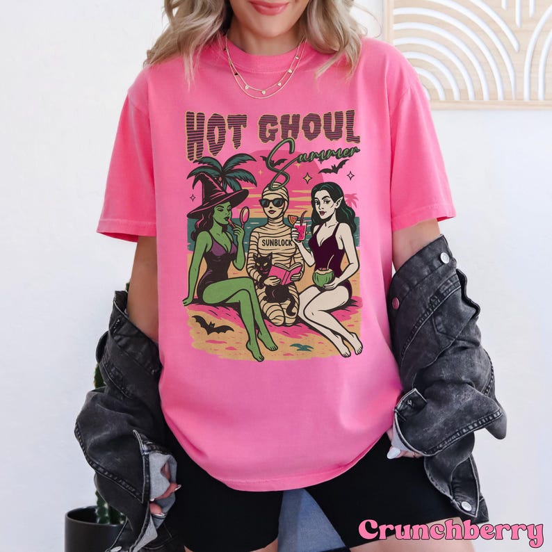 May include: A bright pink t-shirt featuring the phrase "Hot Ghoul Summer" above a graphic of three cartoon figures. The graphic shows a witch, a mummy, and a vampire in swimwear. The witch is wearing a hat and holding a mirror. The mummy is holding a book.