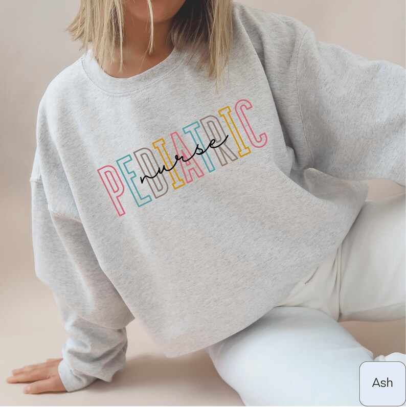 A model wears ash  sweatshirt that says Pediatric nurse. Pediatric is in outlined blocky font. Each letter a different color: melon-pink, med-bright-blue, dark taupe, yellow-gold. Nurse is smaller lowercase script in the middle.