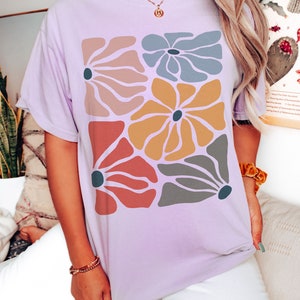 A model wears an orchid shirt with abstract, wavy, boho flowers in colors of muted mauve, light grey-blue, gold, red, and dark taupe all with deep blue pistils. The colorful design is a slightly distressed texture.