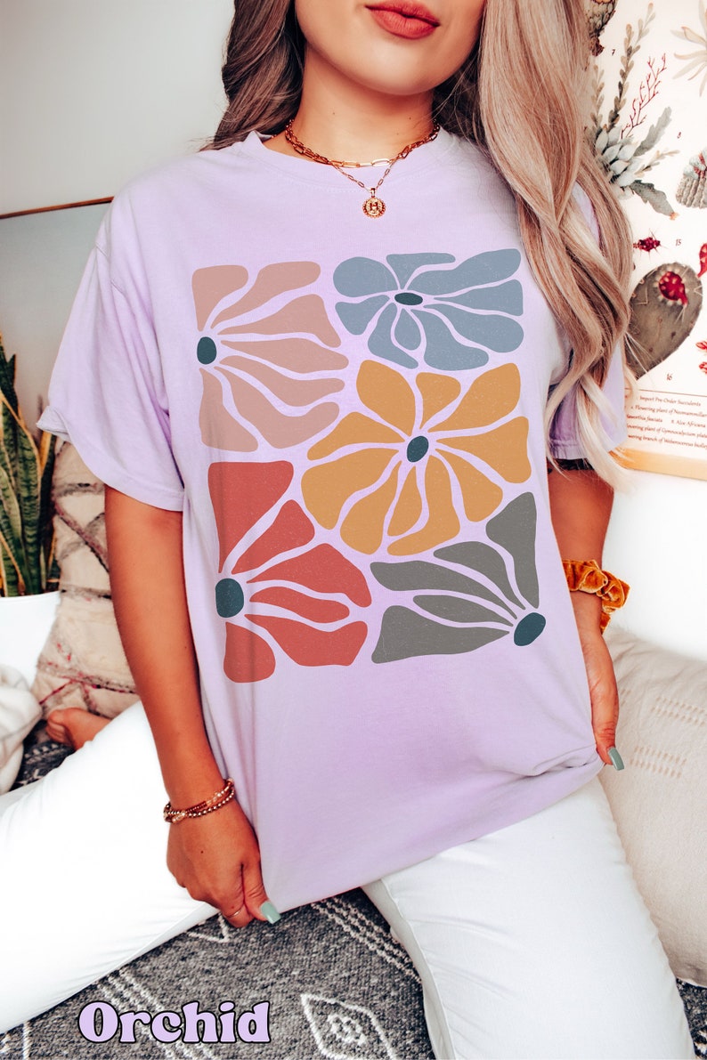 A model wears an orchid shirt with abstract, wavy, boho flowers in colors of muted mauve, light grey-blue, gold, red, and dark taupe all with deep blue pistils. The colorful design is a slightly distressed texture.