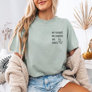 May include: A pale green t-shirt featuring the text "MY FAVORITE INFLUENCERS ARE SAINTS" and a praying hands graphic. The shirt is a loose fit and is worn with denim shorts. The background is neutral with decorative items.