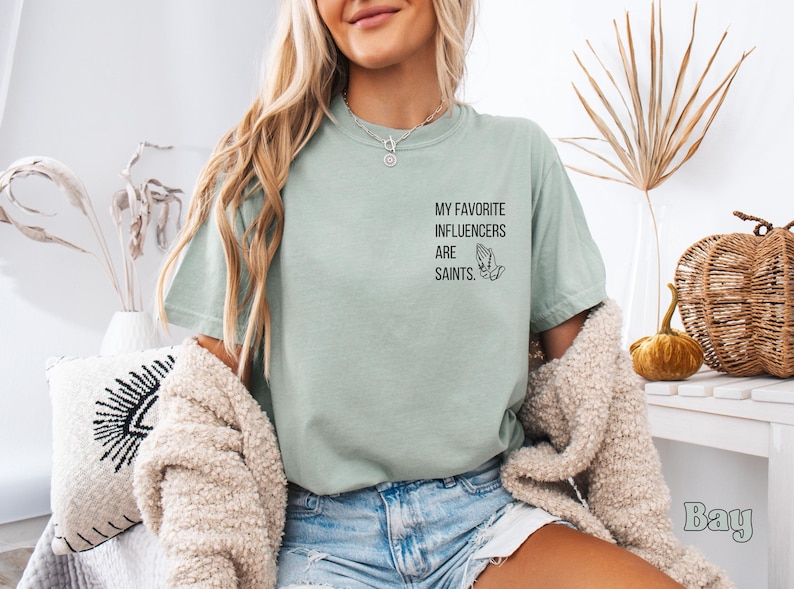 May include: A pale green t-shirt featuring the text "MY FAVORITE INFLUENCERS ARE SAINTS" and a praying hands graphic. The shirt is a loose fit and is worn with denim shorts. The background is neutral with decorative items.