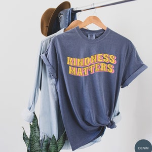 Puede incluir: Una camiseta azul oscuro con el texto "KINDNESS MATTERS" en letras amarillas y rosas. La camiseta est&aacute; colgada en una percha de madera.