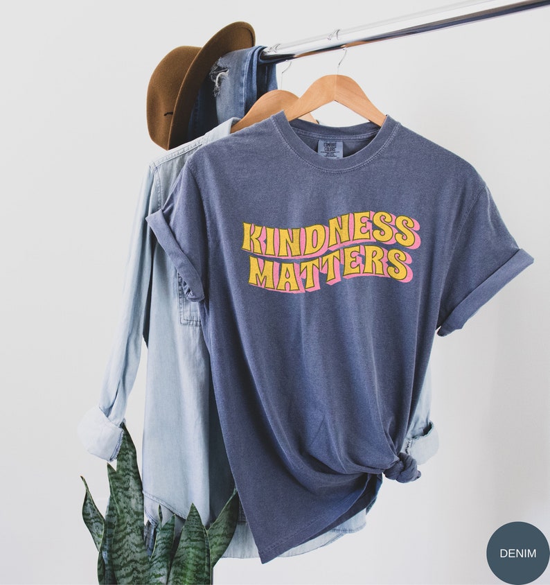 Puede incluir: Una camiseta azul oscuro con el texto "KINDNESS MATTERS" en letras amarillas y rosas. La camiseta est&aacute; colgada en una percha de madera.