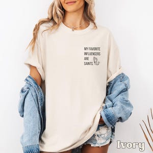 May include: Ivory-coloured t-shirt with the text "MY FAVORITE INFLUENCERS ARE SAINTS" and a praying hands graphic. The shirt has a relaxed fit, with rolled-up sleeves. A light blue denim jacket is draped over the shoulders.
