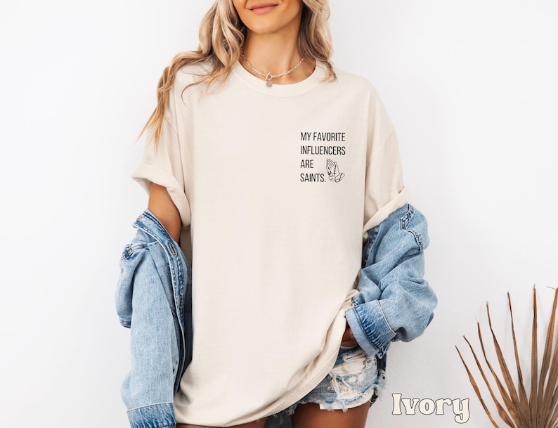 May include: Ivory-coloured t-shirt with the text "MY FAVORITE INFLUENCERS ARE SAINTS" and a praying hands graphic. The shirt has a relaxed fit, with rolled-up sleeves. A light blue denim jacket is draped over the shoulders.