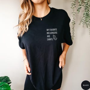 May include: Black t-shirt featuring the white text "MY FAVORITE INFLUENCERS ARE SAINTS." and a graphic of praying hands. The t-shirt has a crew neck design.